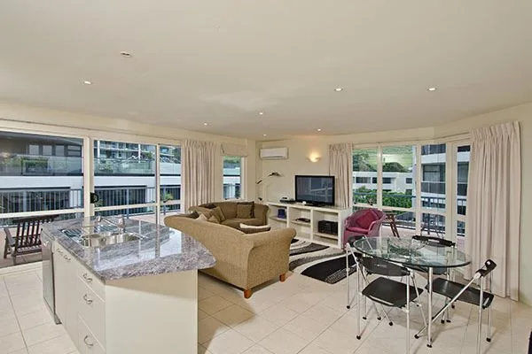 Open-plan living area with kitchen island, sofa, TV, and glass dining table.