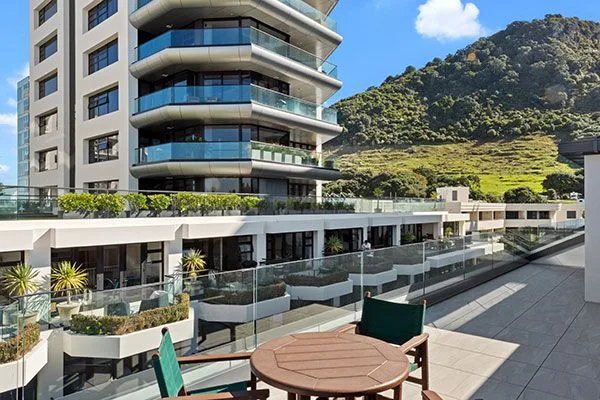 Rooftop terrace with wooden table overlooking a modern building and green mountain.